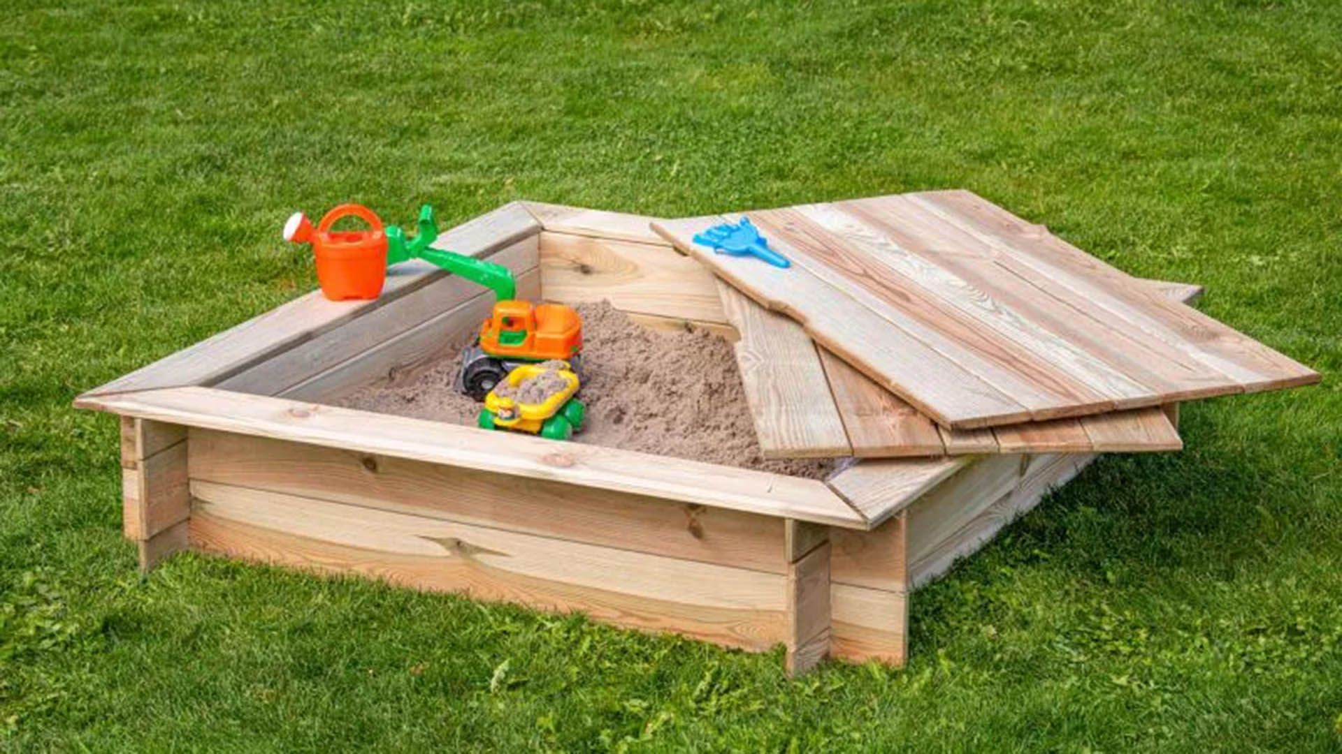 Ein holzumrahmter Sandkasten auf einer grünen Wiese mit farbigem Plastikspielzeug und einem Deckel.