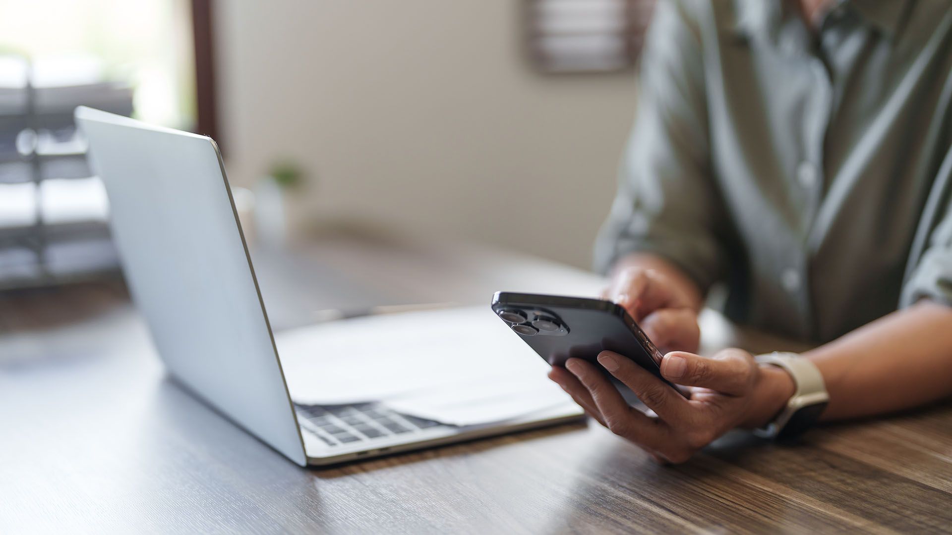 Person benutzt ein Smartphone am Schreibtisch, neben einem offenen Laptop.