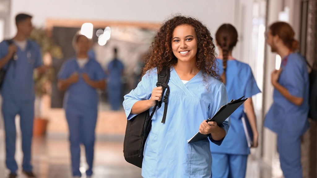Das Bild zeigt eine Gruppe von medizinischen Fachkräften oder Studierenden in blauen Kitteln, die durch einen Flur gehen. Eine Person im Vordergrund trägt einen Rucksack und hält ein Klemmbrett in der Hand. Im Hintergrund sind weitere Personen zu sehen – einige gehen, andere unterhalten sich miteinander. Die Gesichter der Personen sind unkenntlich gemacht.