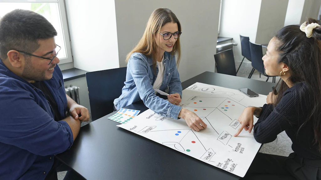 Drei Erwachsene arbeiten gemeinsam an einem Workshop-Poster zu Grundkompetenzen und Weiterbildung.