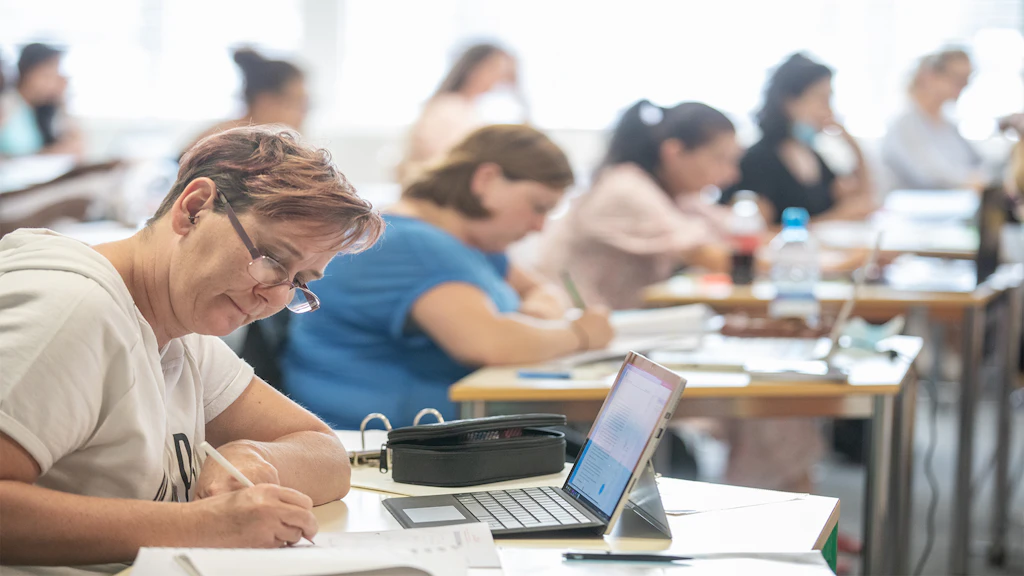 Mehrere Personen sitzen in einem Schulungsraum und schreiben an Tischen, im Vordergrund liegt ein Laptop.
