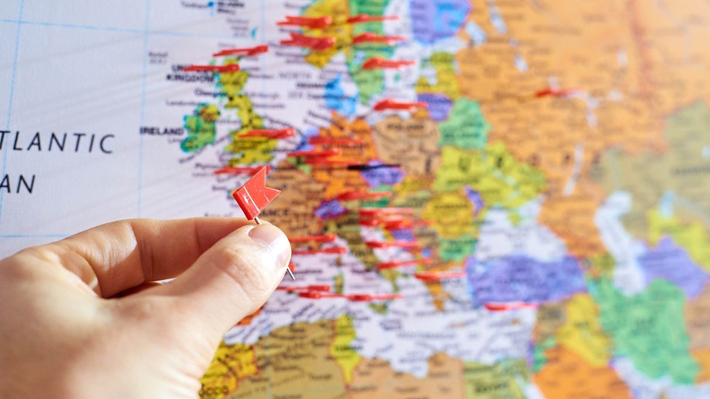 Nahaufnahme einer Hand, die eine kleine rote Stecknadel auf eine Europakarte steckt, auf der bereits mehrere andere rote Stecknadeln verschiedene Orte in Europa markieren.