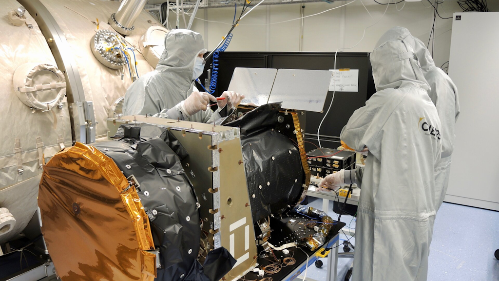 Technical team in cleanroom suits working on a satellite structure covered with metallic and protective foil components.
