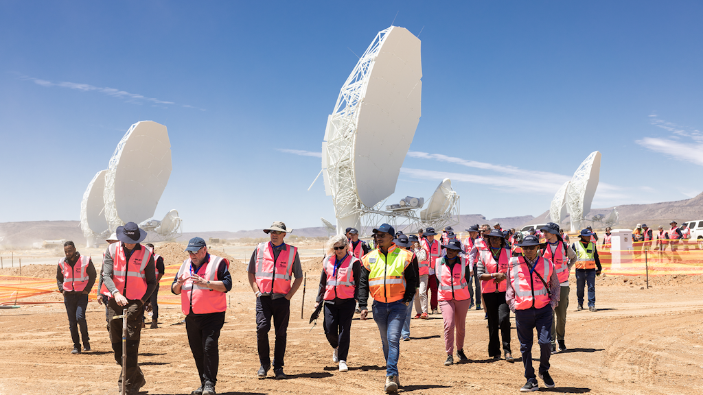 Group of people wearing safety vests walking across a desert site with large radio telescopes in the background.