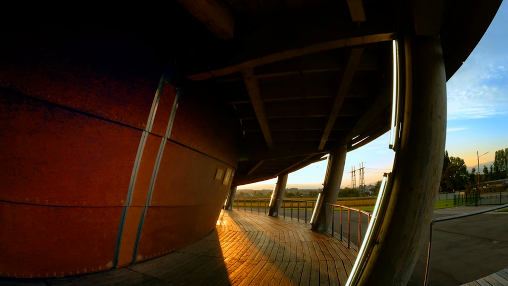 Architectural structure with a curved wooden deck, rounded support pillars, and side sunlight.