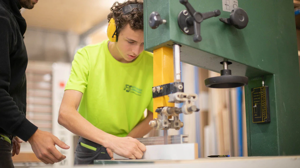 Un jeune homme portant une protection auditive travaille avec concentration sur une scie à ruban dans un atelier, tandis qu’une autre personne à côté de lui lui donne des instructions.