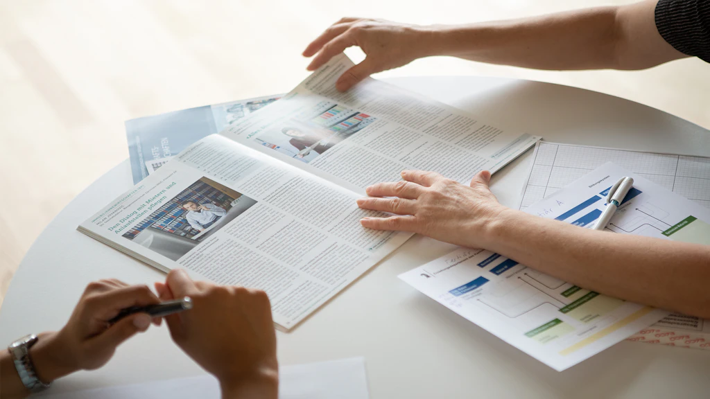 Deux personnes consultent un magazine ouvert et un document avec un diagramme sur une table, une main tourne une page.
