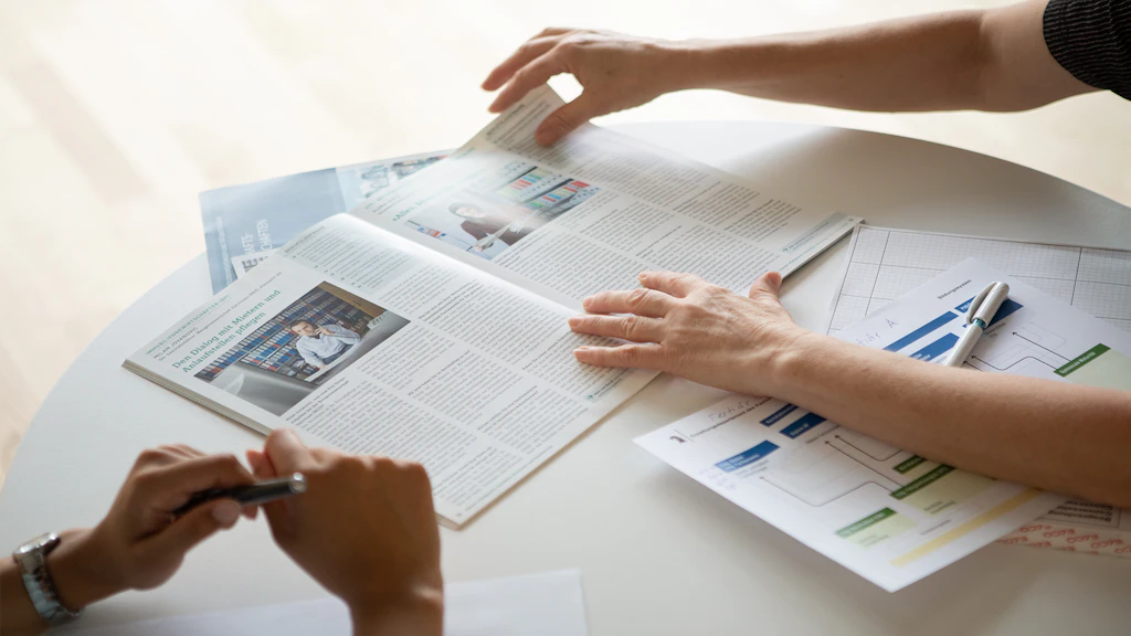 Due persone osservano una rivista aperta e un documento con diagrammi su un tavolo, mentre una mano gira una pagina.
